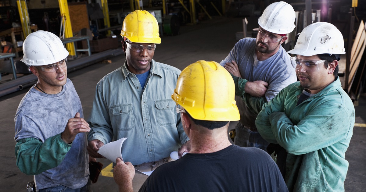 Group of 5 construction workers wearing hard hats talk in a circle