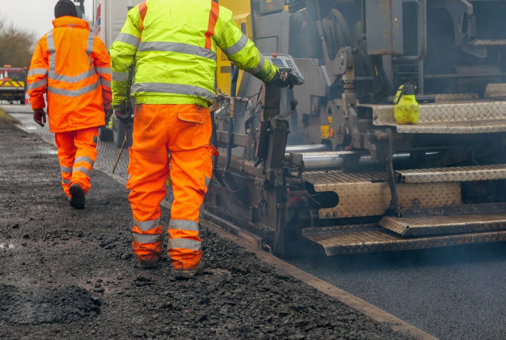 Two construction workers, in high visibility workwear, walking beside an asphalt paver machine which is laying new tarmac on a public road. Using a control panel one of the workmen is controlling the distribution of hot, steaming asphalt concrete, making sure the level of tarmac is consistent. It is slightly compacted, before an asphalt drum compactor follows to roll it further before the new tarmac sets.