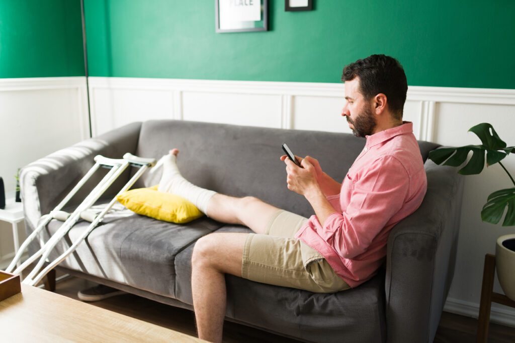 Attractive man with a leg injury texting on his smartphone while resting on the sofa