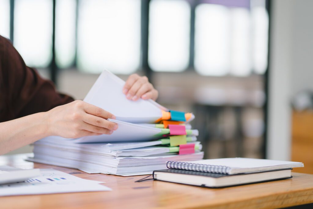The businesswoman's hands are busy working amidst stacks of paper files, searching and checking for unfinished documents among the folders and papers on her cluttered office desk