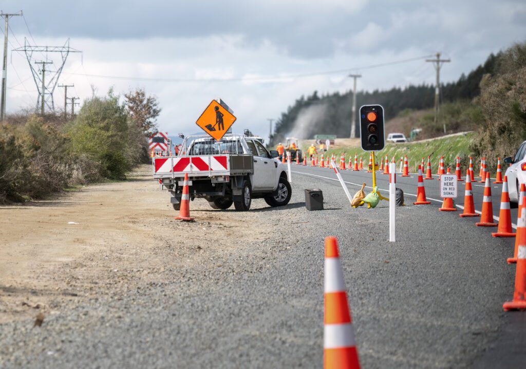 Roadwork sign on top of the work truck. Orange traffic cones on the road. Cars stopped at the roadwork red light. Unrecognizable workers on the road.