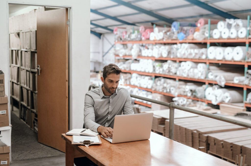 Manager working on a laptop while sitting at his desk in a carpet warehouse with shelves full of stock in the background