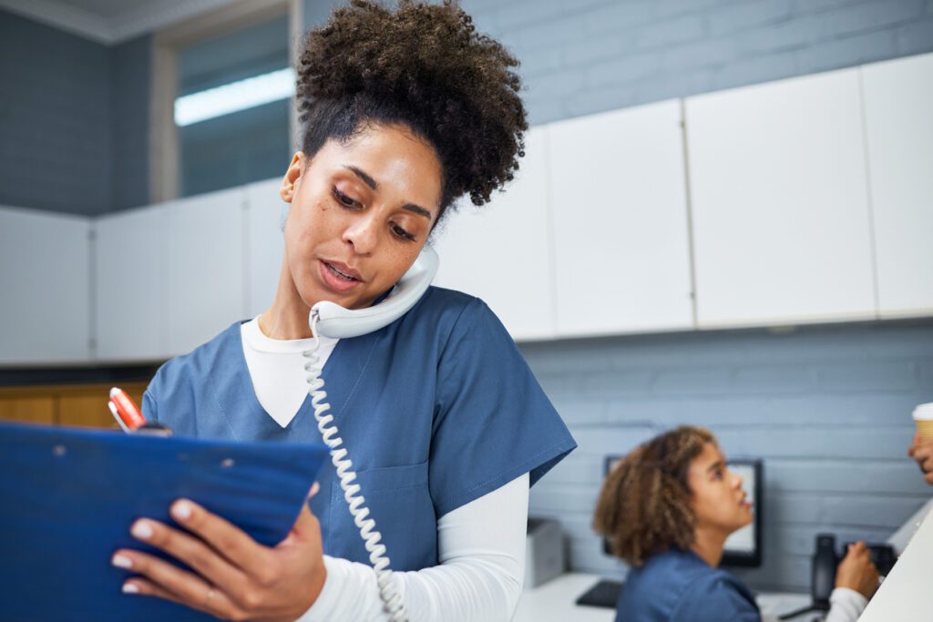 A female healthcare worker multitasks by speaking on the phone and writing notes in a modern medical office setting. She exudes professionalism, interaction, and communication in her administrative duties.