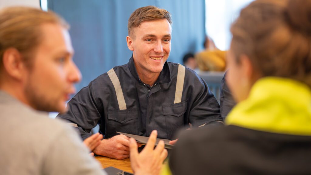 team of oil refinery engineers. wearing in safety wear, one man in casual wear, and another man in workwear discuss something at a table with a laptop. Business meeting, teamwork, and communication concept.