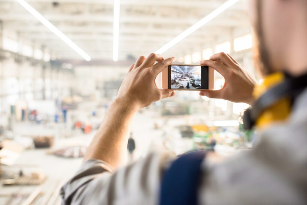 Over shoulder view of unrecognizable inspector wearing ear protectors standing at spacious production department of modern plant and taking picture on smartphone while carrying out inspection.