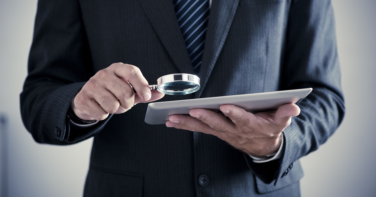 Person in suit examines tablet with magnifying glass